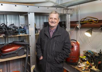 older man with a beard standing inside a tool shed looking at the camera