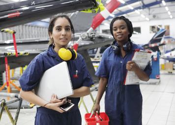 two Black women workers hold helmets and ear protectors standing in front of an airplane 