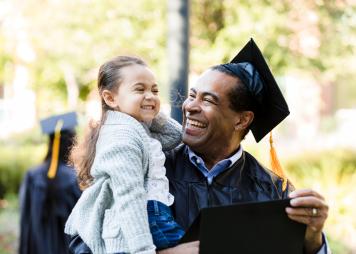 Un hombre con toga y birrete por su graduación sostiene a una niña pequeña en sus brazos; ambos sonríen