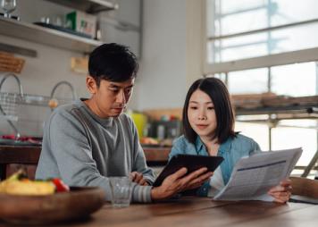 An Asian couple looks at a computer and has papers and receipts on their table