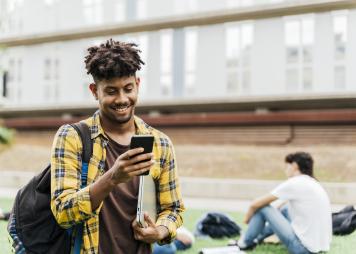 Estudiante universitario caminando en el campus de la universidad, mirando un teléfono inteligente y llevando una computadora portátil.