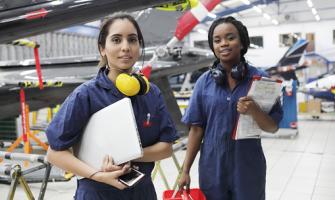 dos trabajadoras negras sostienen cascos y protectores auditivos paradas junto a un avión