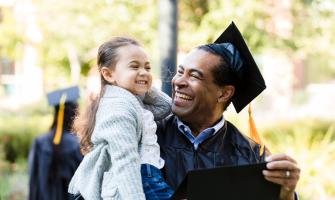 Un hombre con toga y birrete por su graduación sostiene a una niña pequeña en sus brazos; ambos sonríen