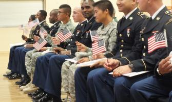Una fila de hombres sentados con uniformes militares sosteniendo pequeñas banderas de EE. UU. a la espera de prestar juramento