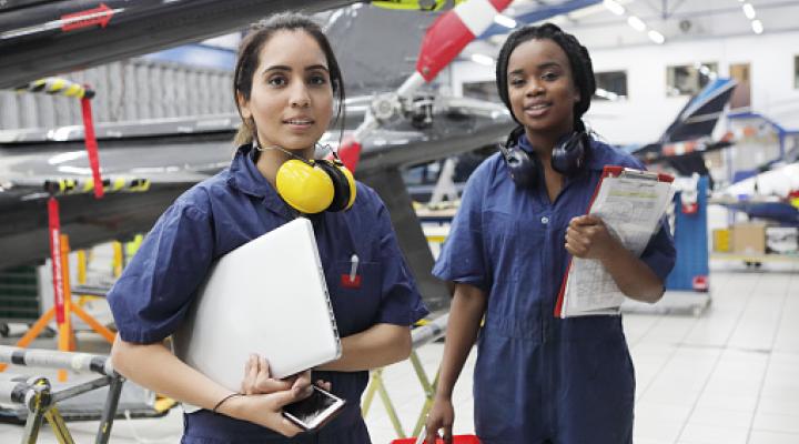 two Black women workers hold helmets and ear protectors standing in front of an airplane 