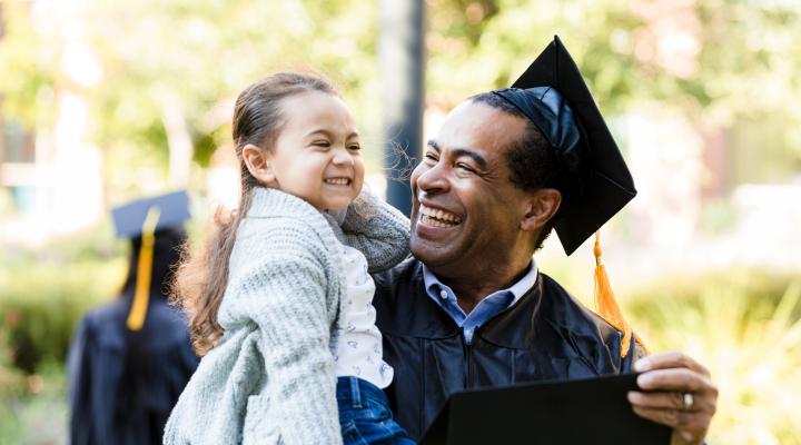 Un hombre con toga y birrete por su graduación sostiene a una niña pequeña en sus brazos; ambos sonríen