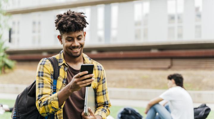 College student on college campus walking and looking at smart phone holding laptop.